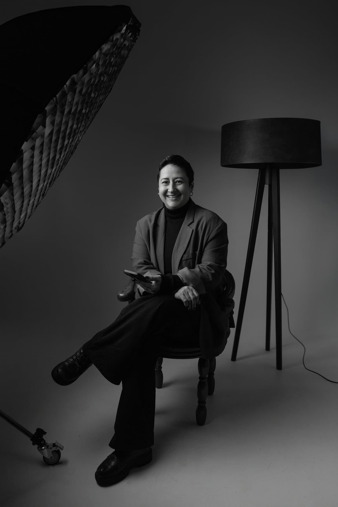 Evelyn Rodrigues sentada no estúdio, entre o softbox e a luminária, sorrindo com o celular na mão